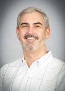 Portrait of Charles Schambeau smiling against a gray background.