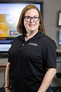 UCF Professor Claire Connolly Knox, wearing glasses and a black polo shirt, smiles for the camera, with a TV screen softly blurred in the background.