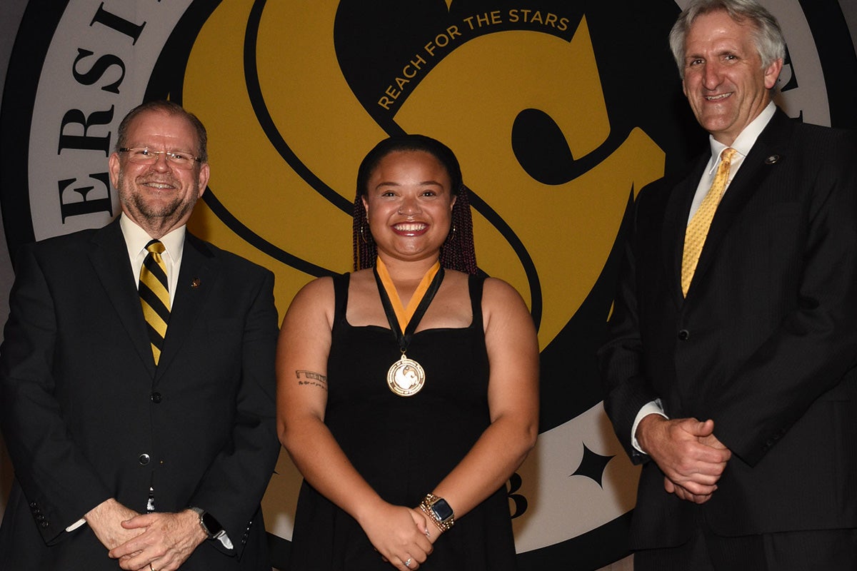 UCF President Alexander N. Cartwright and Provost John Buckwalter stand with a student wearing an Order of Pegasus medal, with a large Pegasus seal displayed in the background.