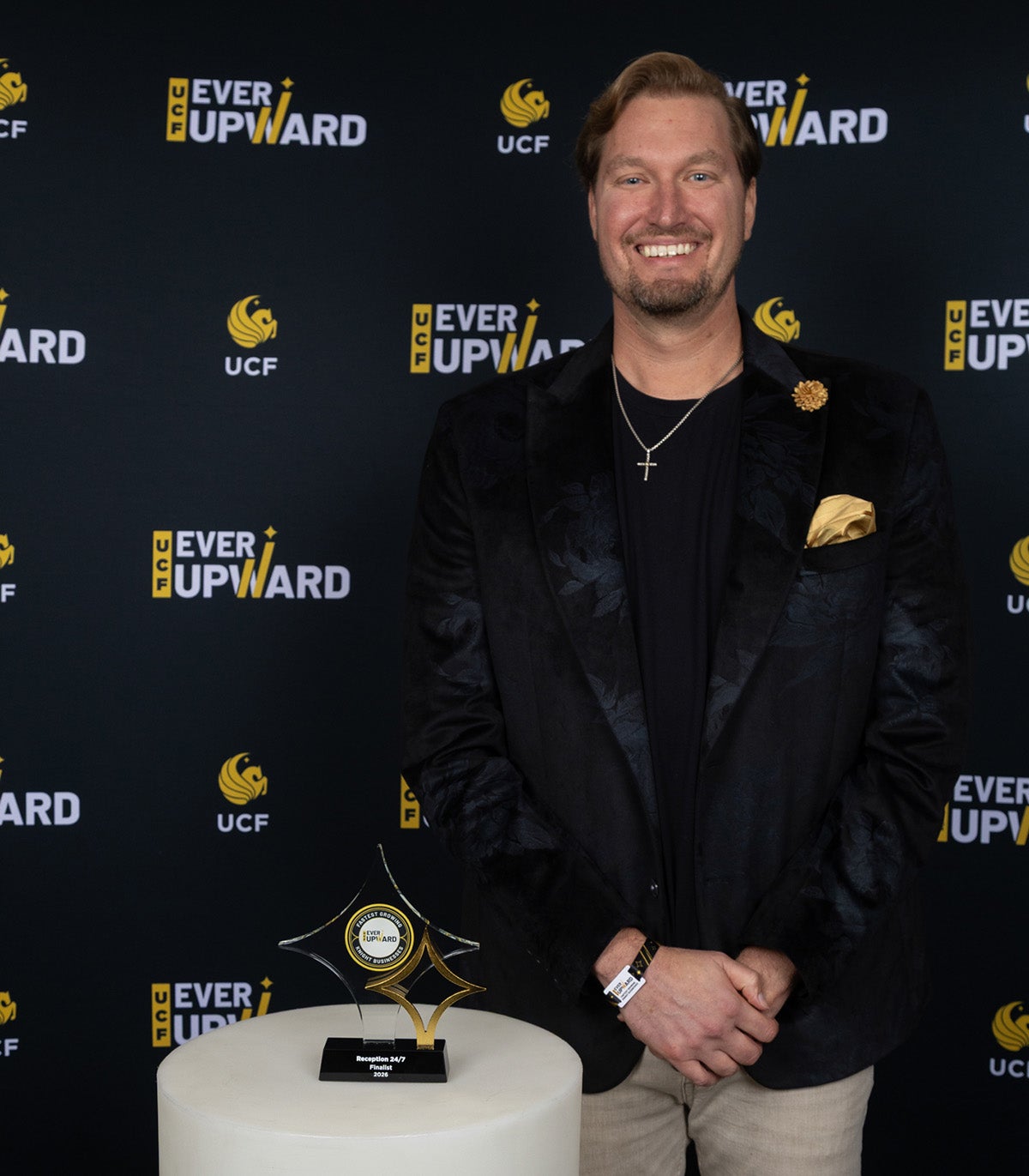 Blonde, bearded man in black business suit and tan pants stands next to white cylinder table with glass award on top