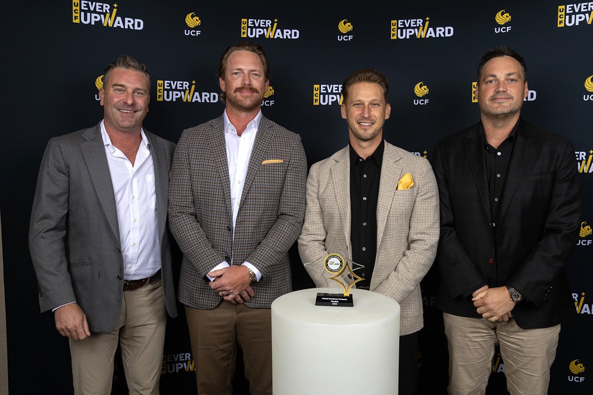 Four men in business suits stand side by side behind white cylinder table with glass award on top
