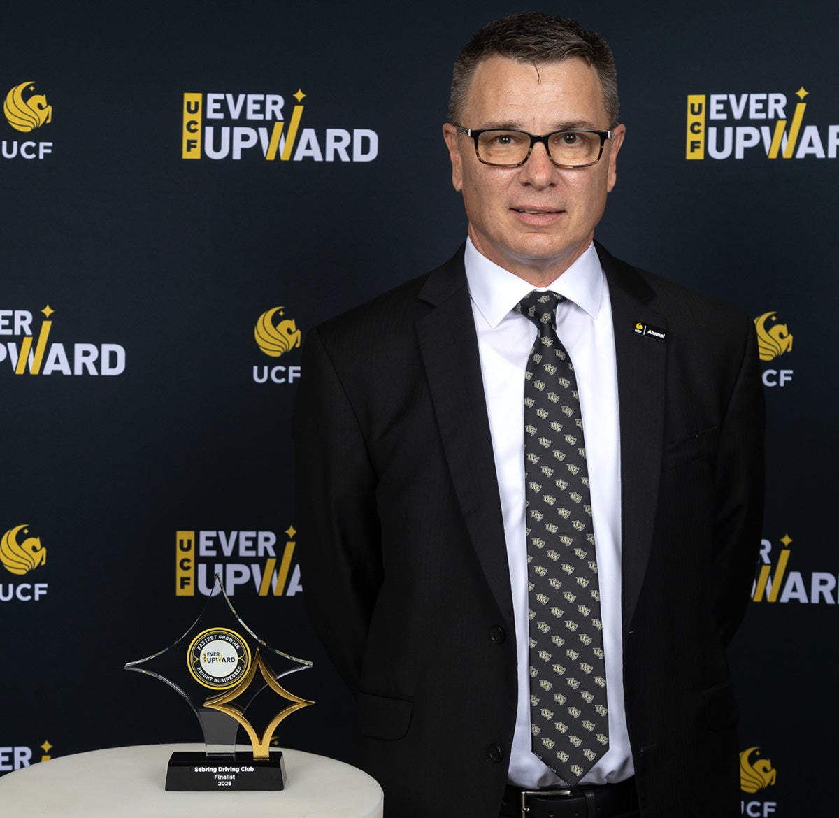 Man wearing glasses in black business jacket, white shirt, and black and white polka dot tie, stands next to white cylinder table with glass award on top