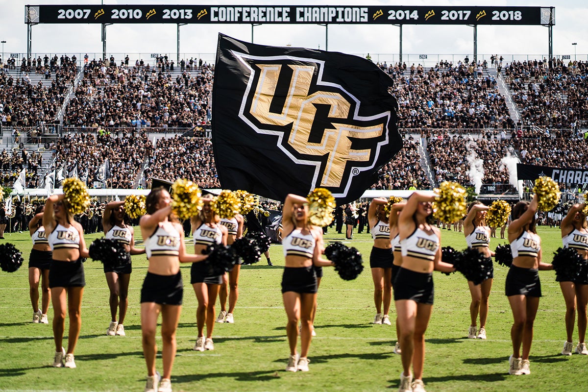 UCF cheerleaders hold black and gold pompoms in front of black and gold UCF flag on football field