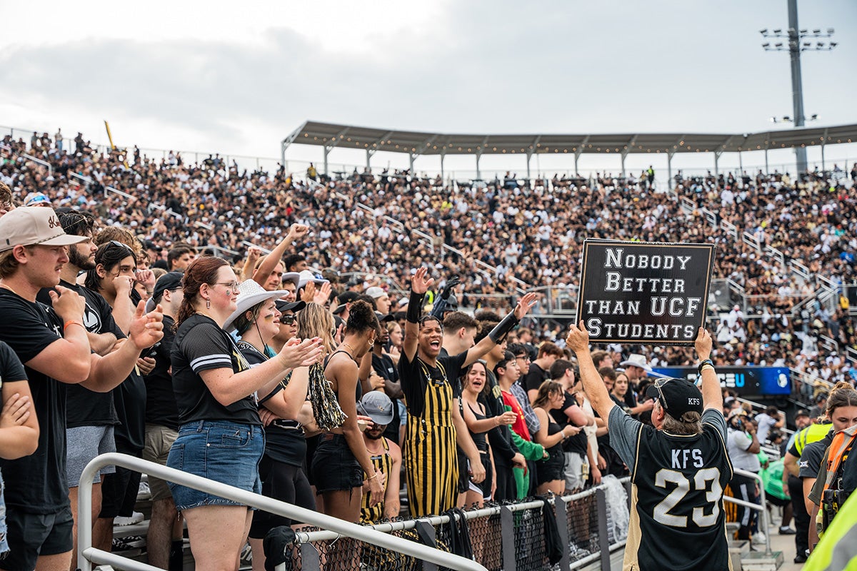 Man wearing UCF jersey holds sign that reads "Nobody better than UCF students" while standing on concourse in stadium in front of rows of students 