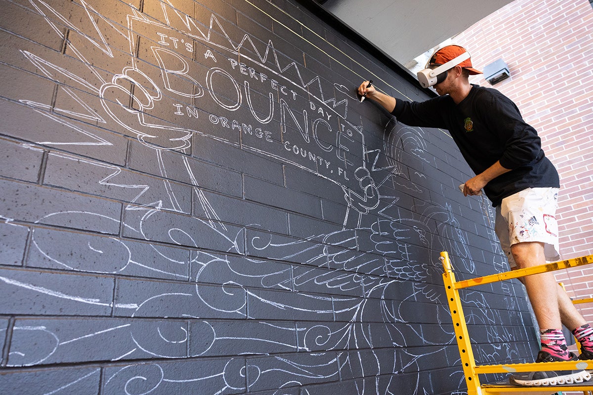Man wearing backwards baseball hat, long-sleeve back shirt and white cargo shorts, wearing VR headset stands on yellow scaffolding as he applies white paint to black wall.