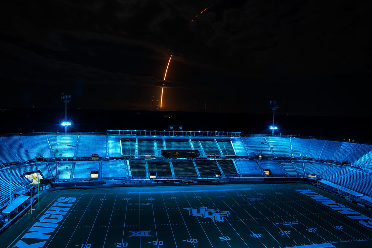 UCF's football field and stands illuminated in blue lighting at night with an orange rocket flair streaking across the sky