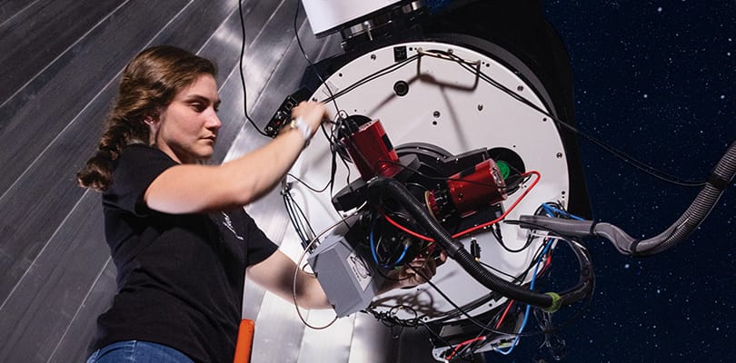 UCF student adjusts equipment on a large telescope at night, surrounded by cables and machinery, with a starry sky in the background.