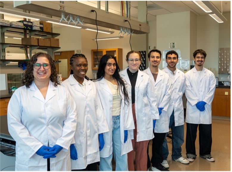 UCF Assistant Professor Cali Calarco stands with six researchers wearing lab coats in a biomedical sciences laboratory.