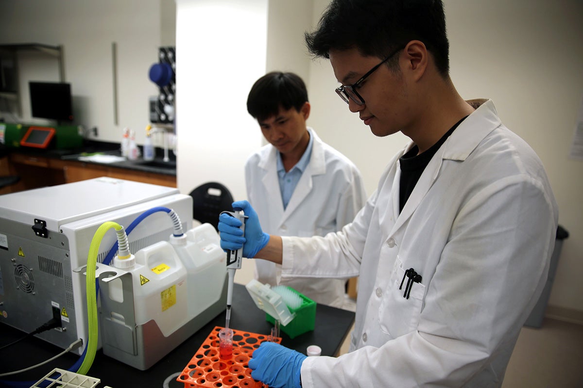 Man in white lab coat, wearing glasses, holds dropper with red liquid in right hand as another man in lab coat observes.