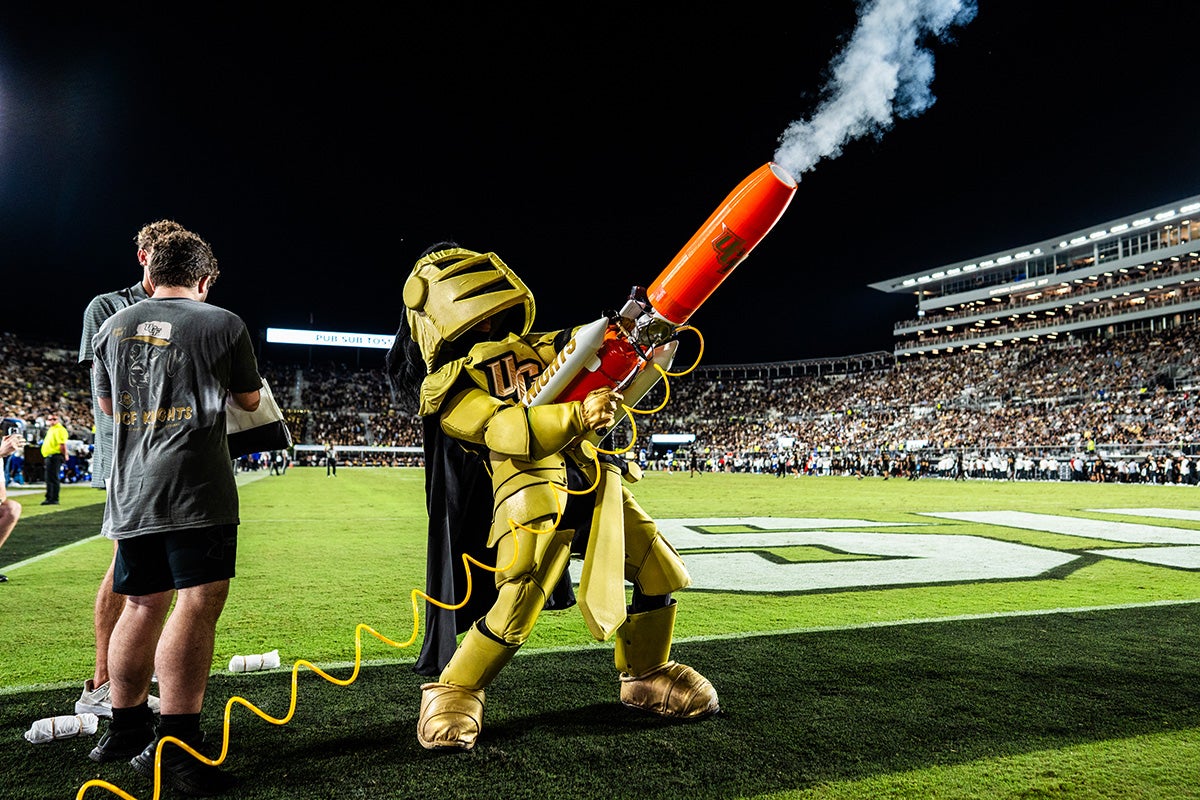 UCF mascot Knightro stands near end zone of football field at night while holding an orange t shirt launcher with white smoke billowing from the cannon