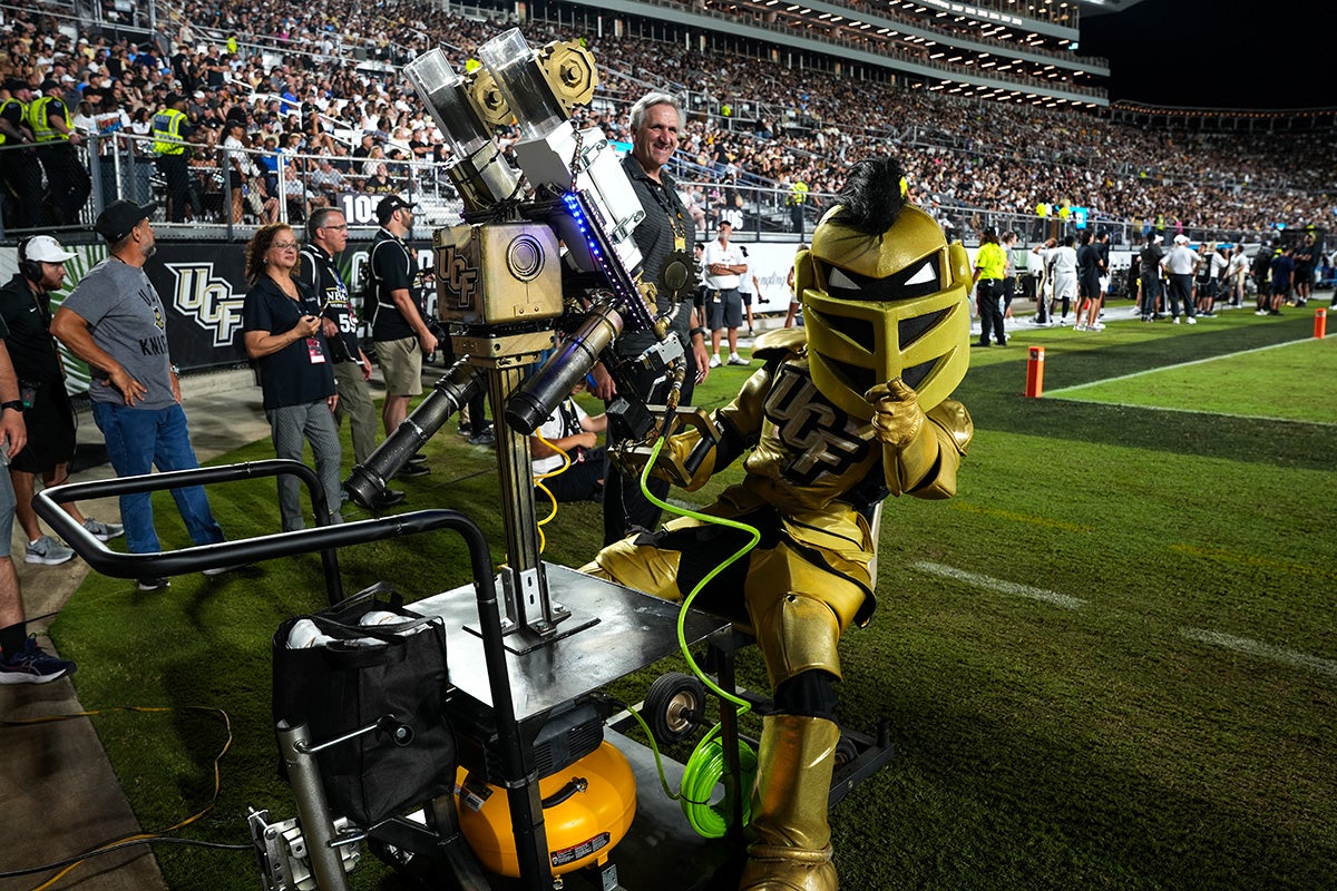 UCF mascot Knightro points to camera while leaning back on seat next to T shirt cannon on UCF football field
