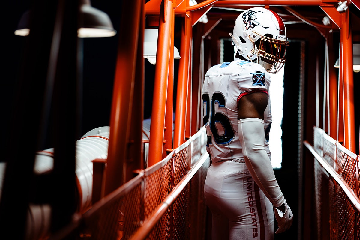 UCF football player in white uniform and helmet looks over his right shoulder as back faces the camera while standing on orange scaffolding