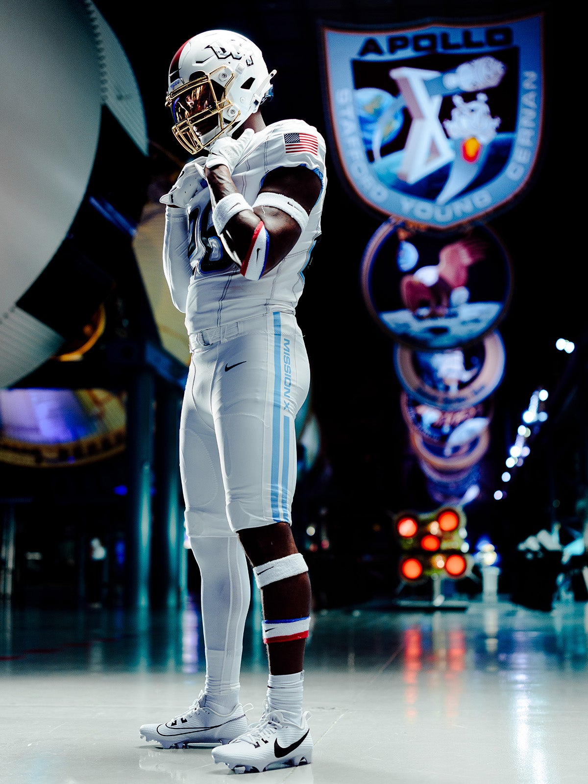 UCF football player in white helmet and white uniform with red, white and blue USA accents stands in atrium featuring Mission patch banners hanging from rafters