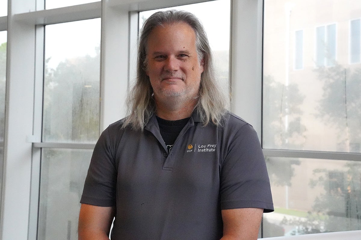 Bearded man with shoulder length gray hair wearing a gray polo shirt stands in front of glass windows with steel beams