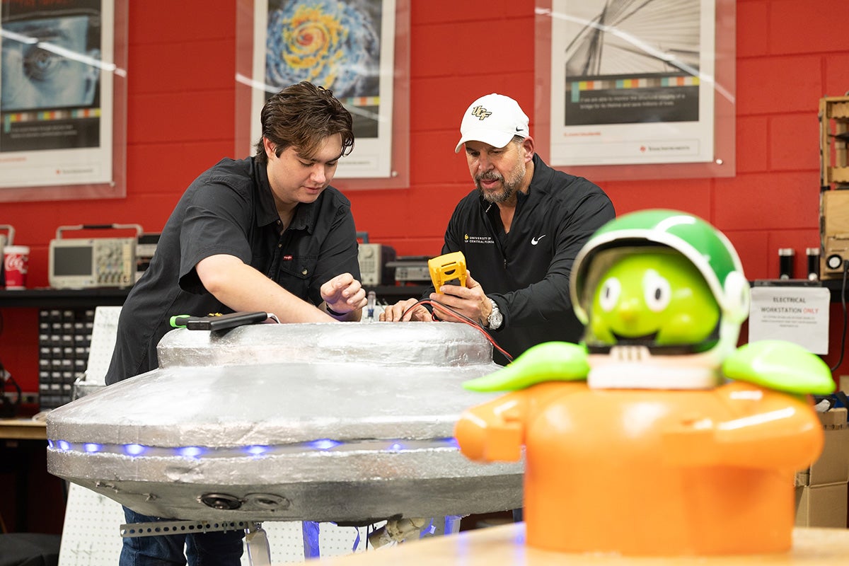 College-aged male with brown hair and another man wearing white UCF baseball hat tinker with silver saucer shapeship in engineering lab with Citronaut figure on lab bench next to them