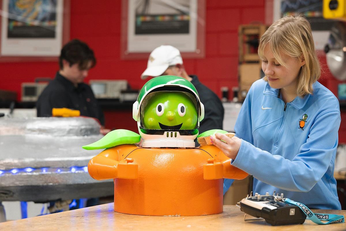 Blonde college-age woman wearing blue Citronaut long sleeve shirt paints figure of Citronaut on lab bench
