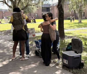 Pair of women hug male and female college student with open coolers next to sidewalk outside under trees