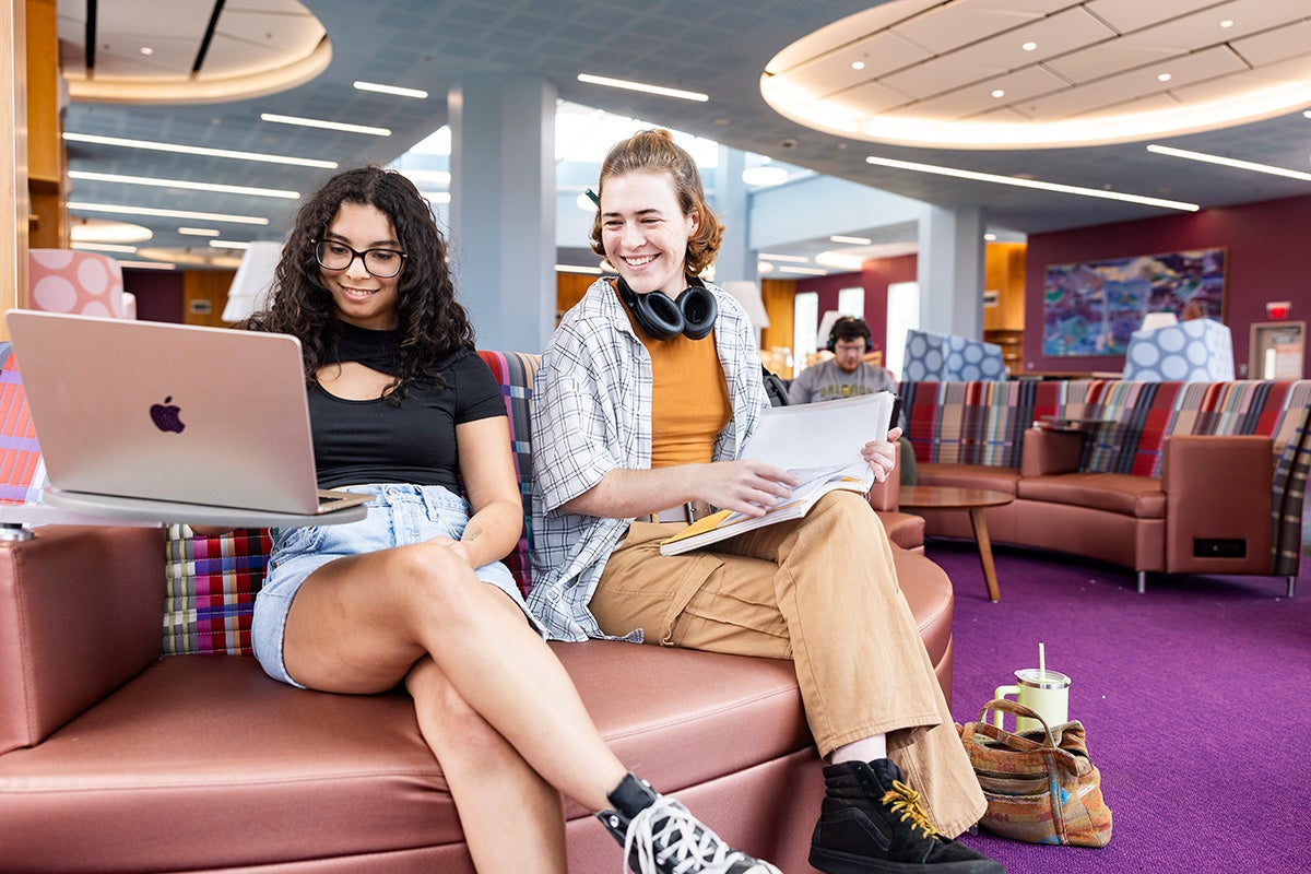 two women sit on rose-colored sofa with laptops open in library