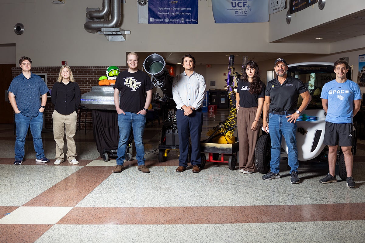 Six UCF students and facility manager Pete Alfieris stand in front of Citronaut spaceship, Knightro's golf cart, bat signal and t shirt launcher in atrium of Engineering building
