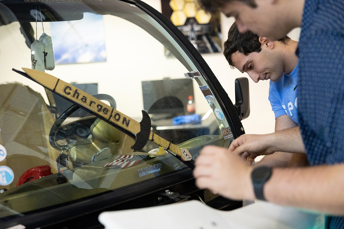 Two college males hover near windshield of golf cart with a "charge on" sword wiper blade