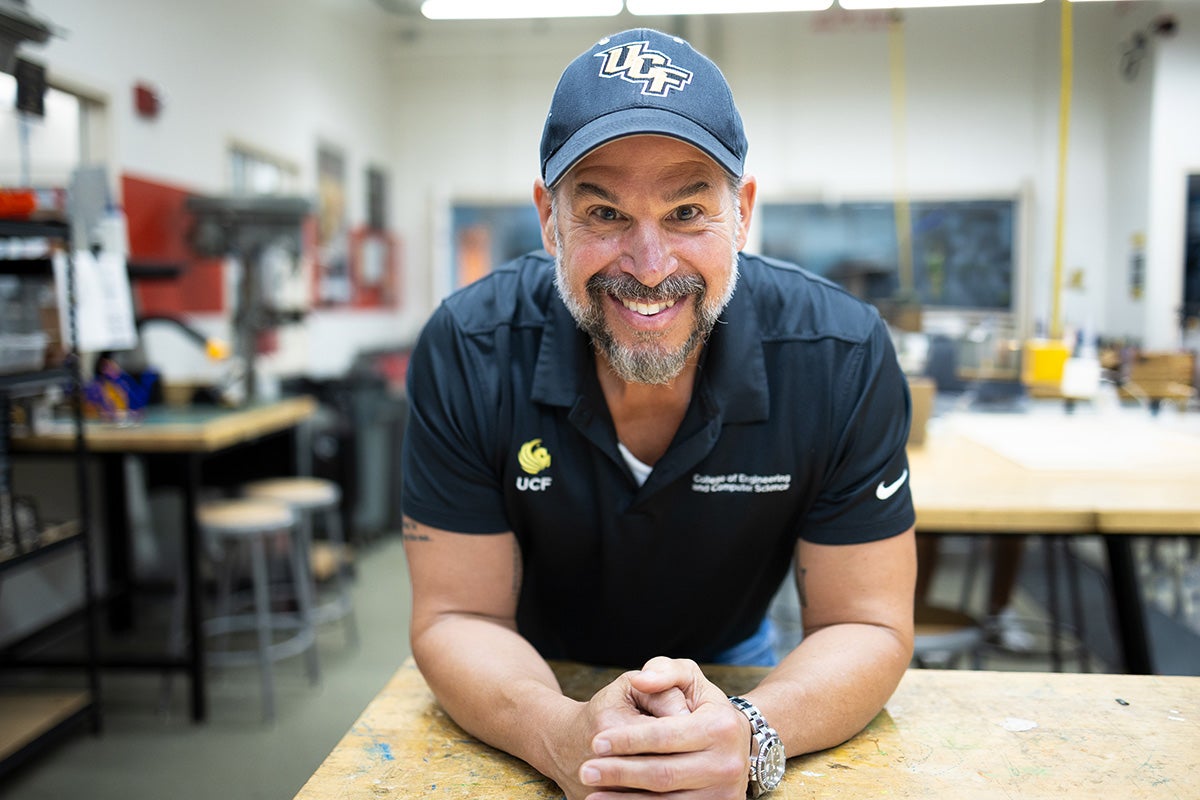 Bearded man in UCF baseball hat and UCF black polo leans forearms on wood desk in lab