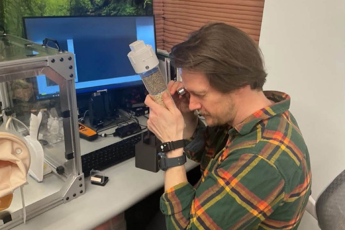 Man with brown hair wearing green and orange plaid shirt holds magnifying class to eye as he examines small pebbles in a glass container