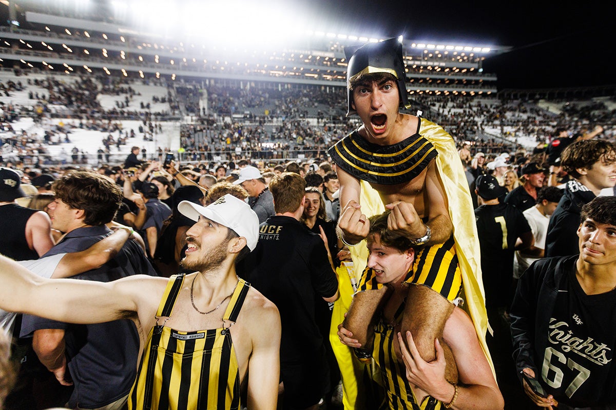 Student wearing black helmet and gold cape rides the shoulders of another UCF fan surrounded by fans on the field at the Bounce House under the stadium lights