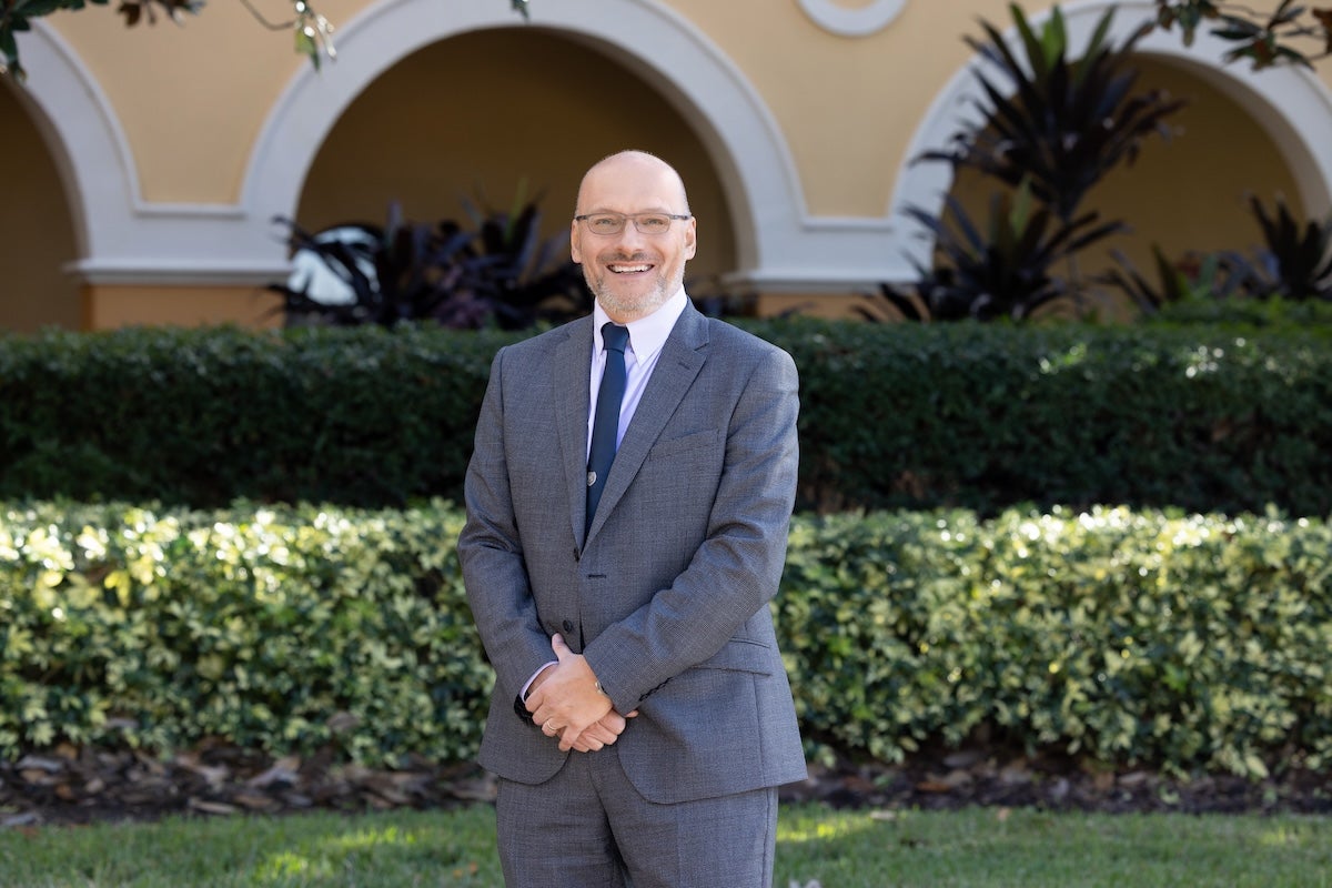Visit Orlando Endowed Chair of Tourism Marketing Alan Fyall poses in a grey suit outside a yellow building with arched walkways.