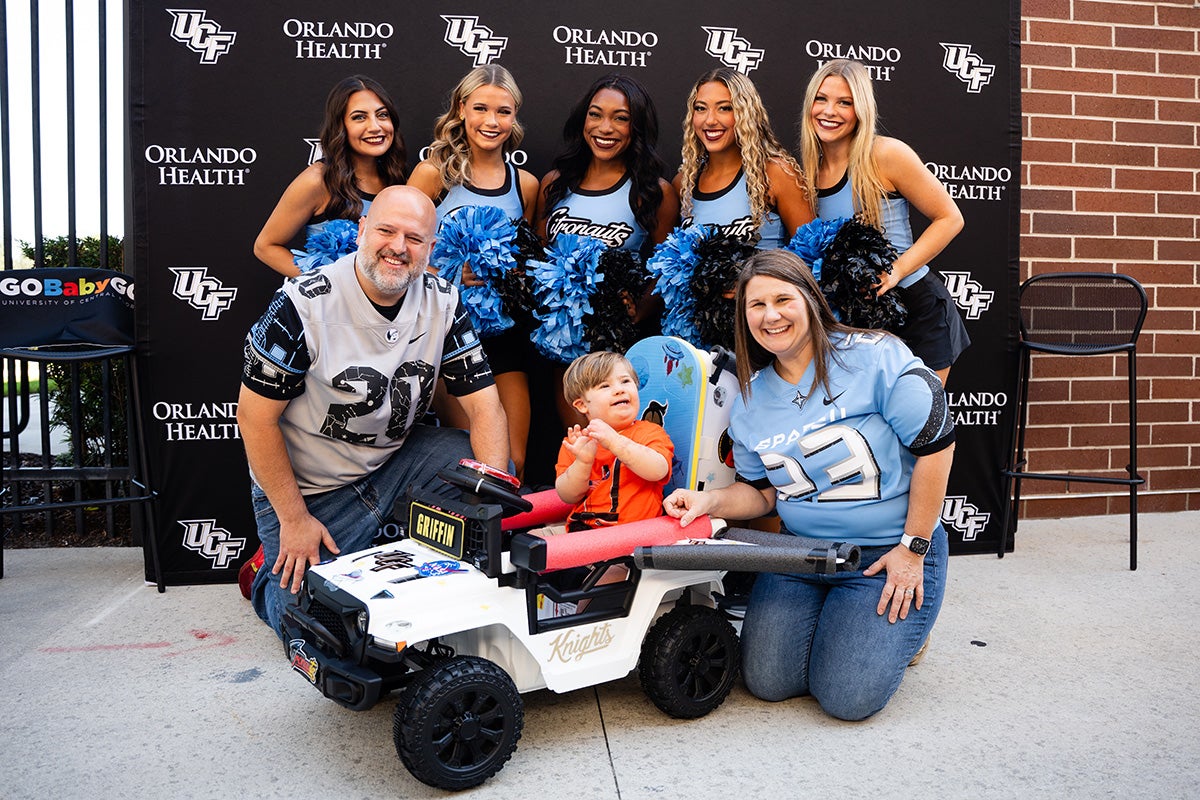 Five UCF dance team members in blue Citronaunt uniforms pose for photo with 2-year-old blonde boy in white motorized toy car with his parents kneeling next to him.