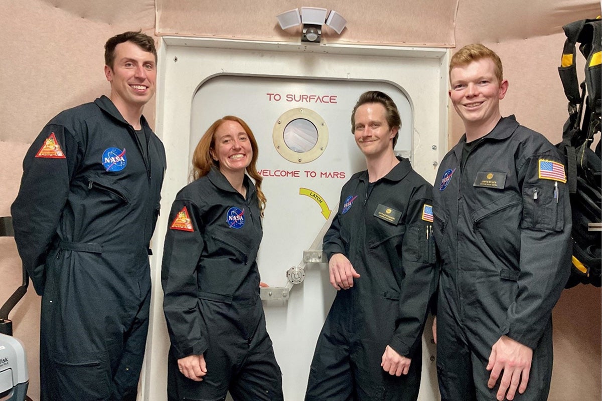 three men and one woman in NASA jumpsuits stand in front of white air lock door that reads "To Surface. Welcome to Mars" in red letters