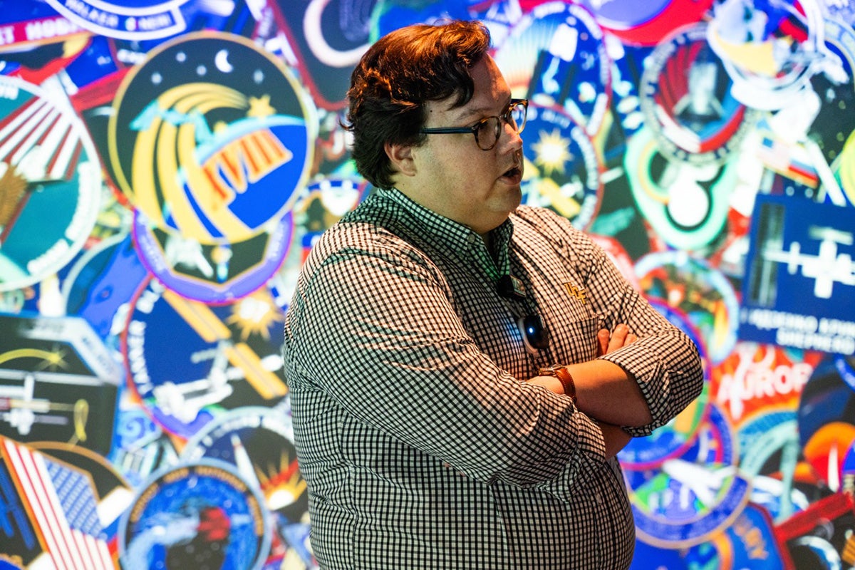 Side profile of man with arms crossed in front of colorful wall with stacked mission space patches