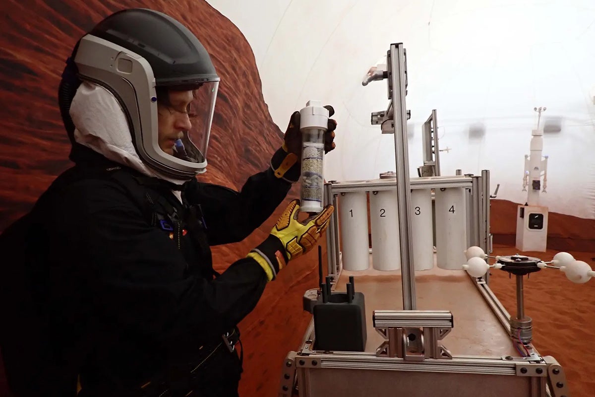 Man wearing black jumpsuit and helmet holds cylinder shaped bottle with red rock and white sky in background
