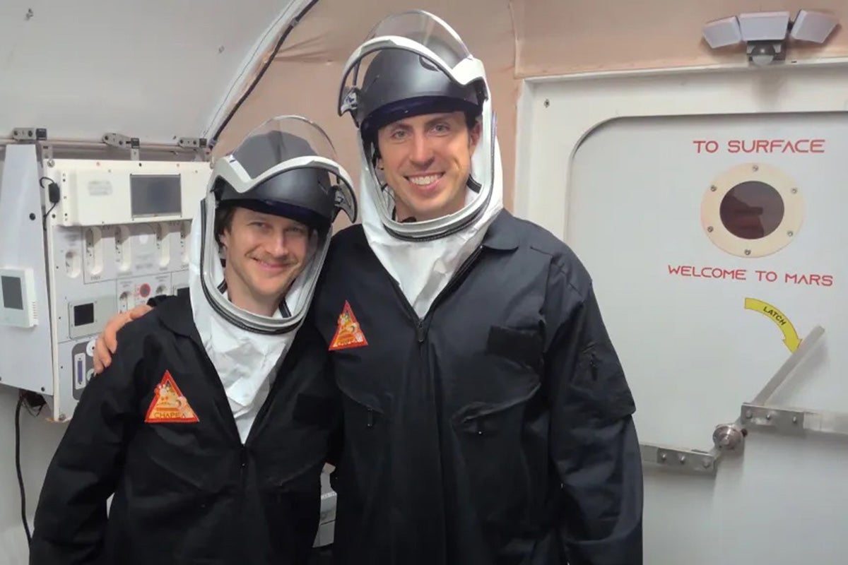 Two men in space suits and helmets pose in front of airlock door that reads To Surface, Welcome to Mars