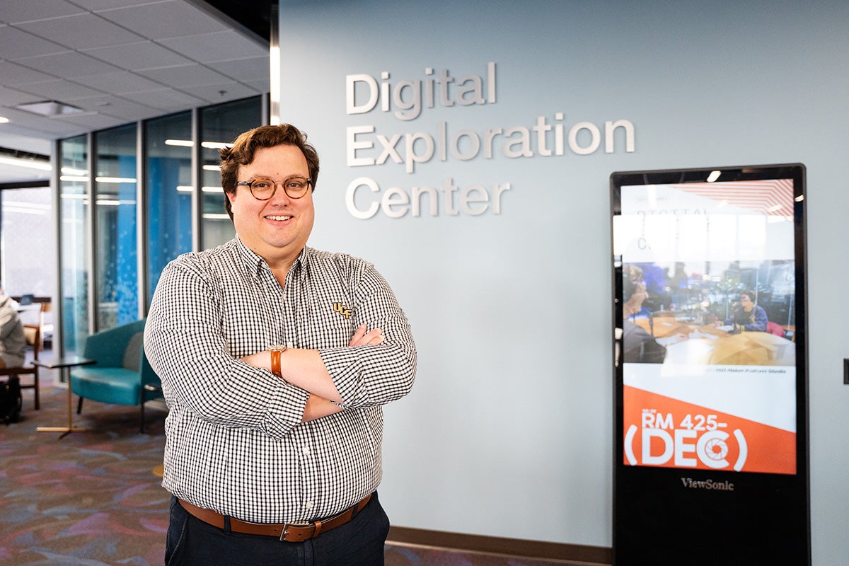 Man wearing glasses and longsleeve collar shirt stands with arms crossed in front of light blue wall with signage that reads Digital Exploration Center