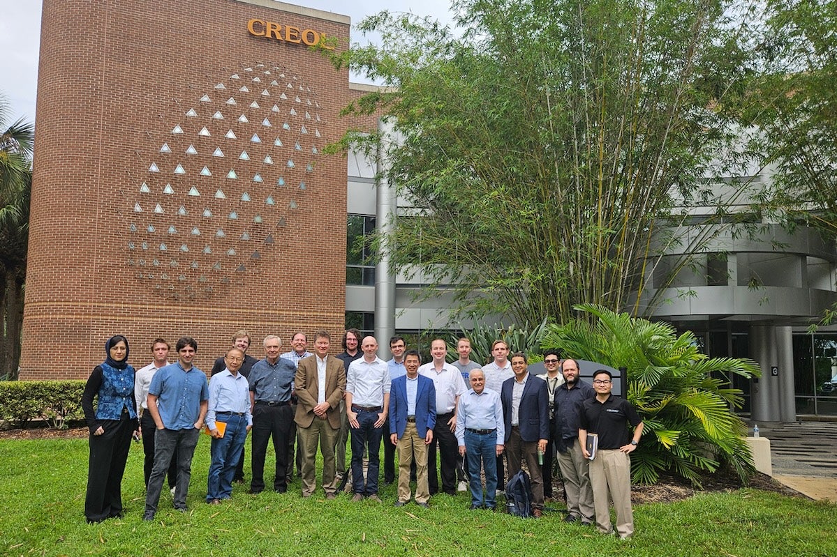 A group of researchers and colleagues pose for a photograph outside UCF’s College of Optics and Photonics building, with the CREOL sign visible in the background.