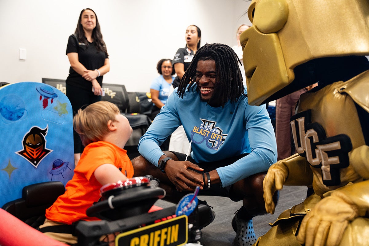 UCF football player in longsleeve dry-fit blue shirt kneels and smiles next to toddler boy and Knightro