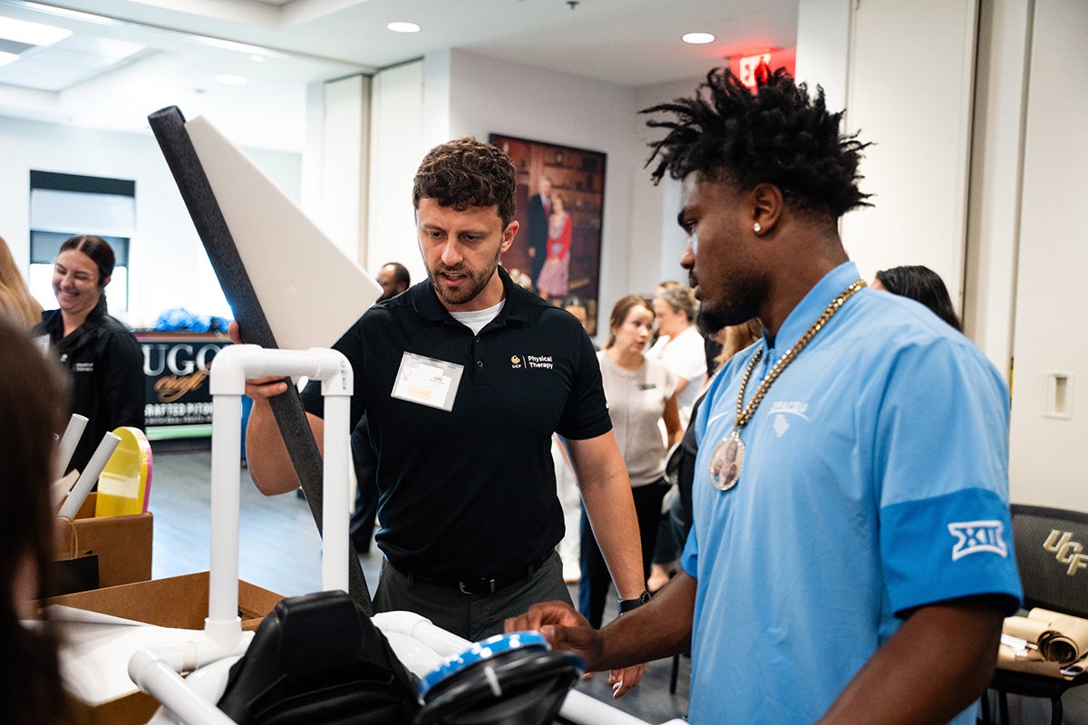 A physical therapy male student in black polo t shirt holds a triangular shaped piece of white foam board as a UCF football player in a blue polo shirt and chain around his next assists