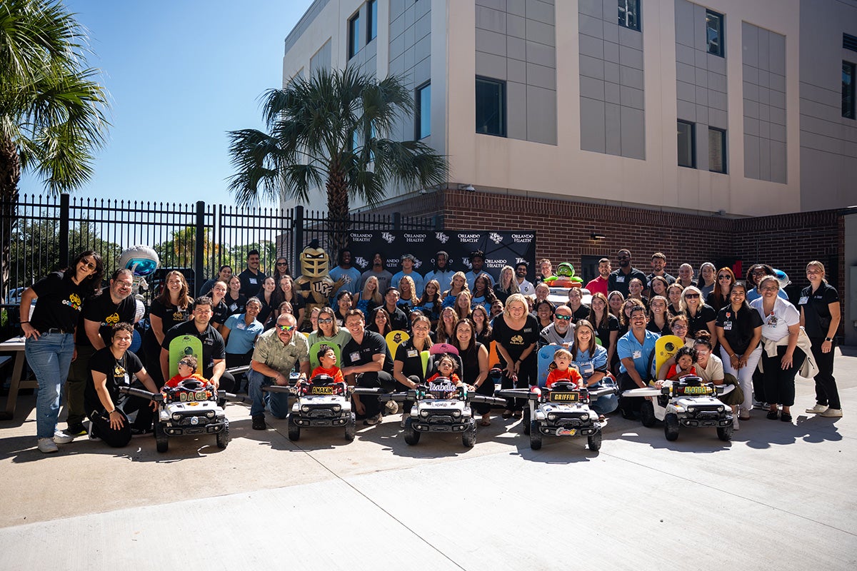 A large group photo with five children in toy motorized cars front and center outside the Wayne Densch Center for Student-Athlete Leadership