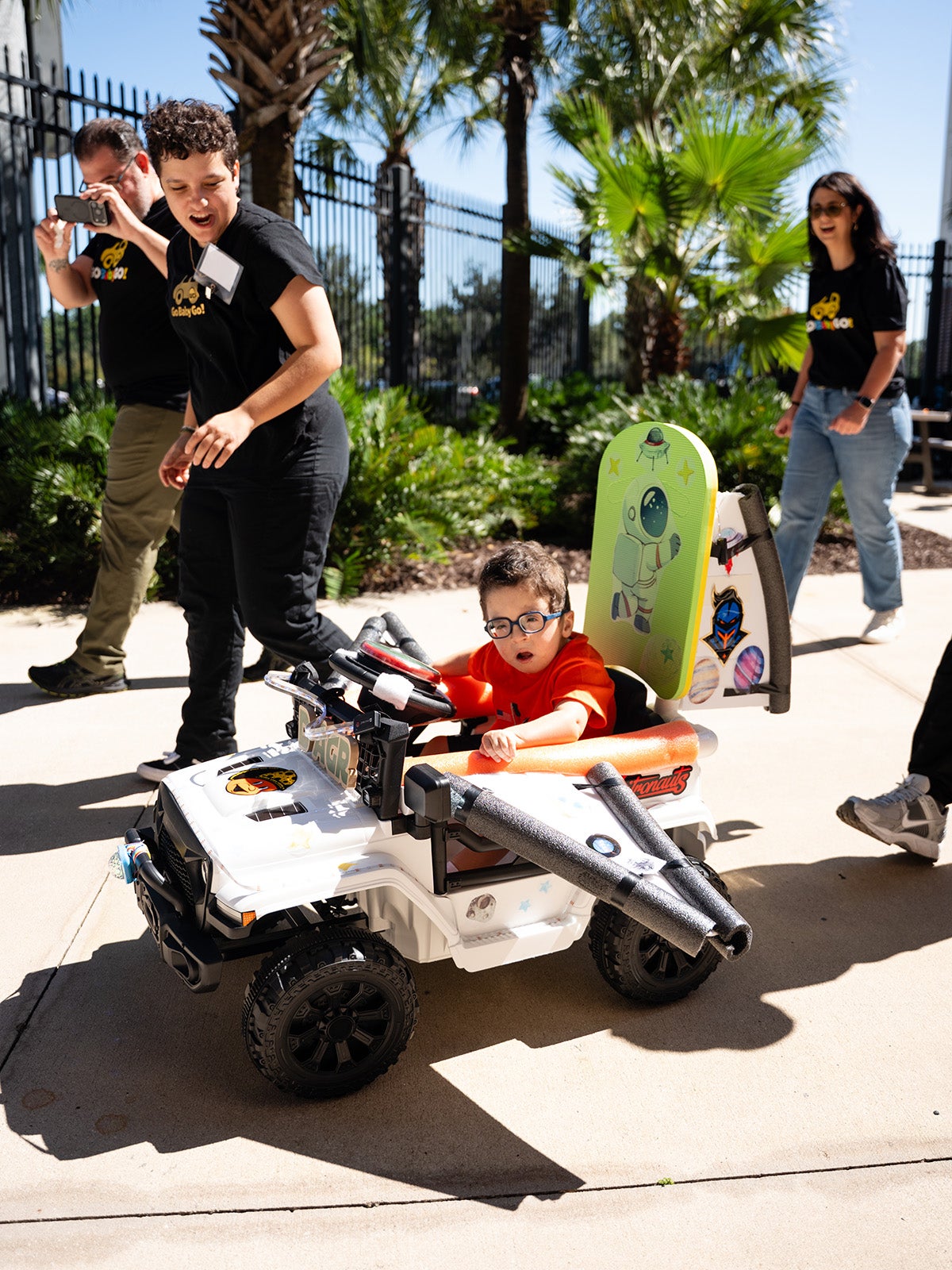 Young boy in motorized toy car as woman in black t shirt and black pants walks alongside him.