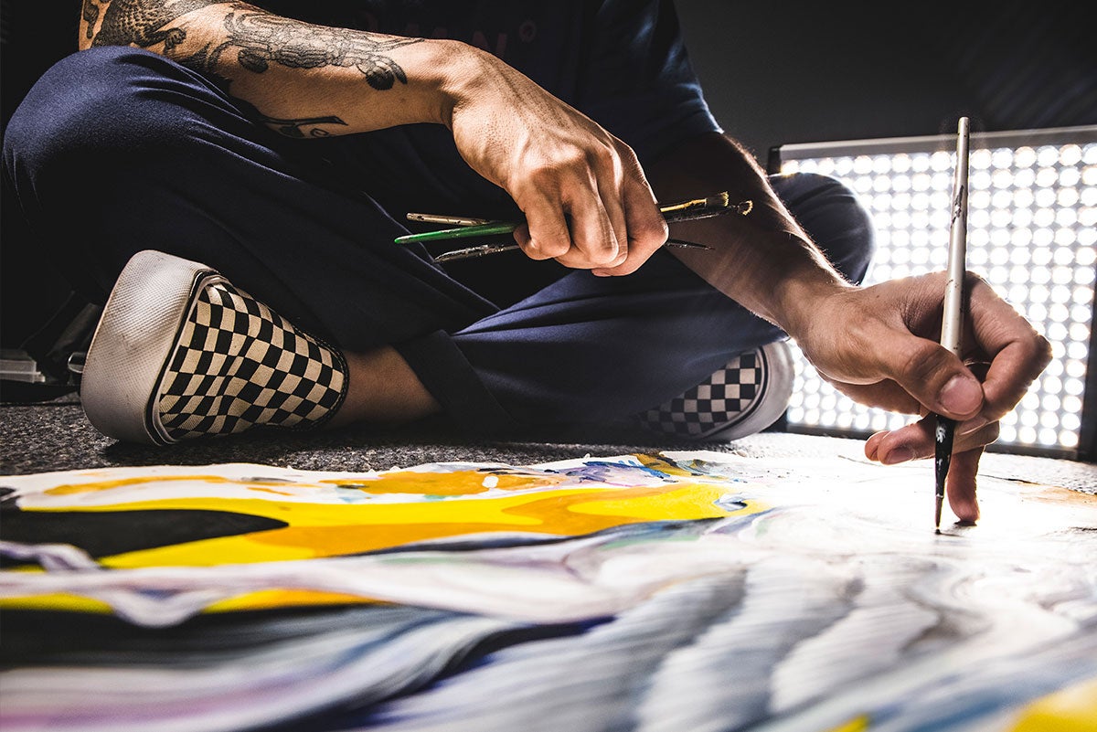 Boy Kong sits on the floor and paints a draft of what the mural at UCF will look like. A bright light box is behind him.