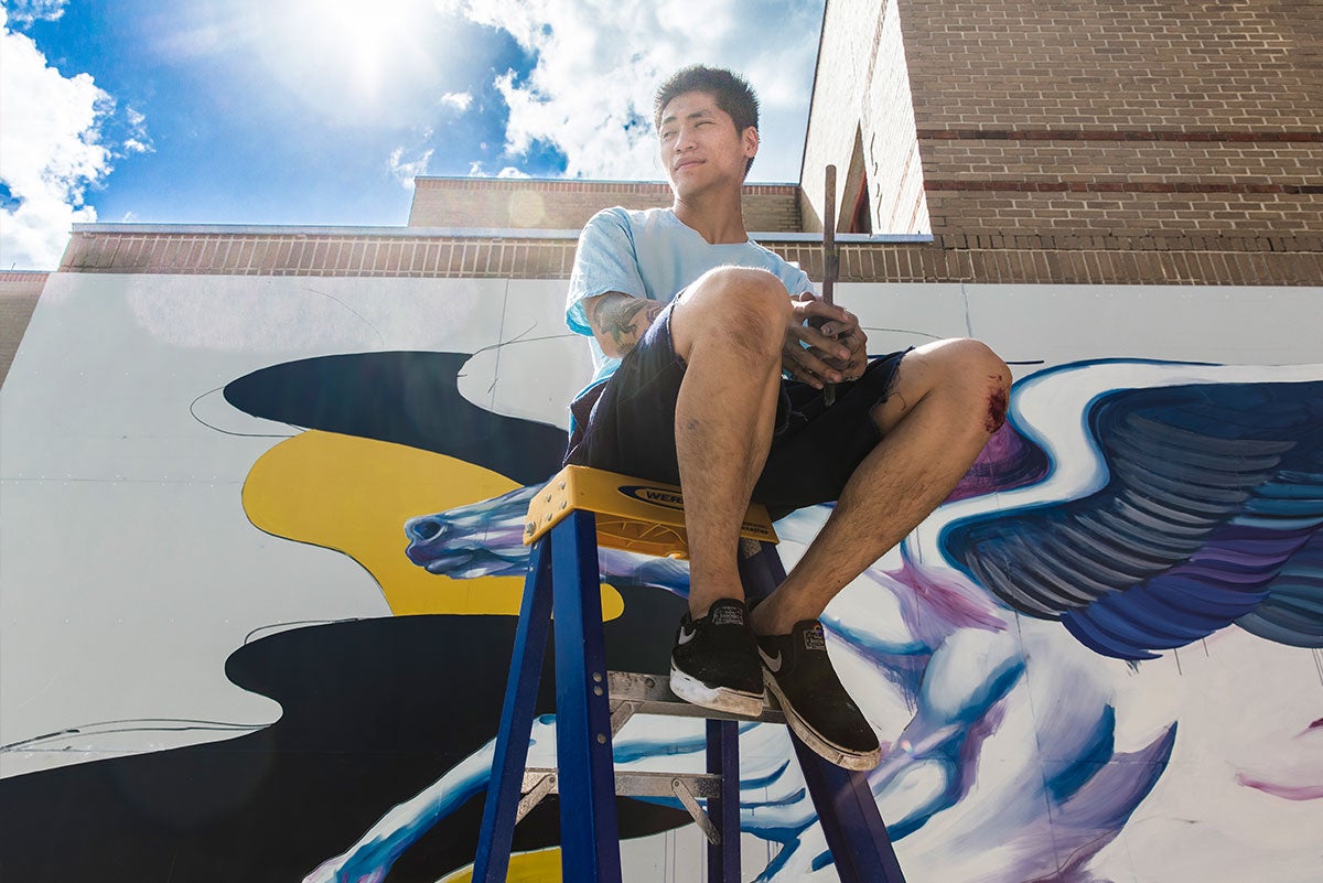 Boy Kong sits on the top step of a ladder holding a paint brush. In the background is the Pegasus mural at UCF in progress. The light from the sun glares in the top of the frame with a bright blue sky with white clouds.