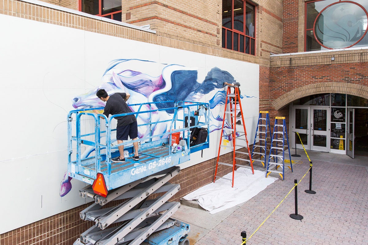 Standing on a blue lift, Boy Kong paints details on the face of Pegasus that is still a work in progress while his brother Chris Troung stands on an orange ladder and fills in the black background that will be a wing. Part of the entrance to the Student Union is in view.