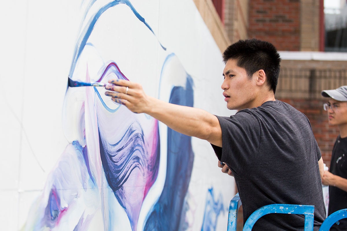 Boy Kong paints the outline of a wing on a large white background on a wall.
