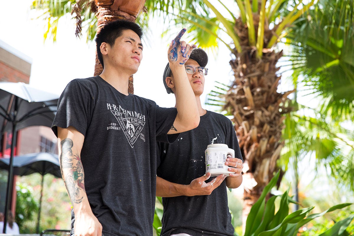 Artist Boy Kong points his finger up at something out of frame. His brother Chris Troung holds a paint can. A palm tree is in the background.