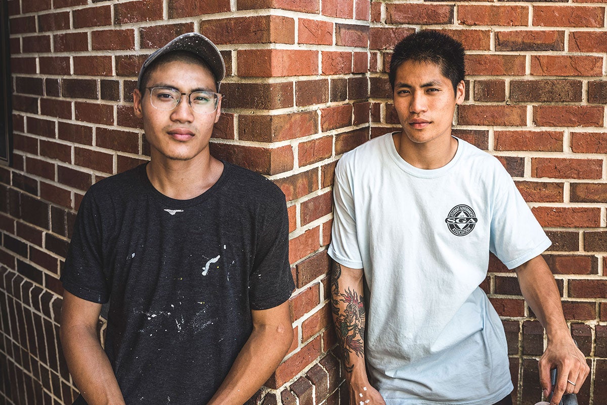 Chris Troung in a black shirt with paint on it is wearing glasses and a tan hat. His brother, artist Boy Kong, stands to the right of him, in a light blue shirt. His tattoo on his left arm is visible. They are leaning against a brick wall and looking directly into the camera.