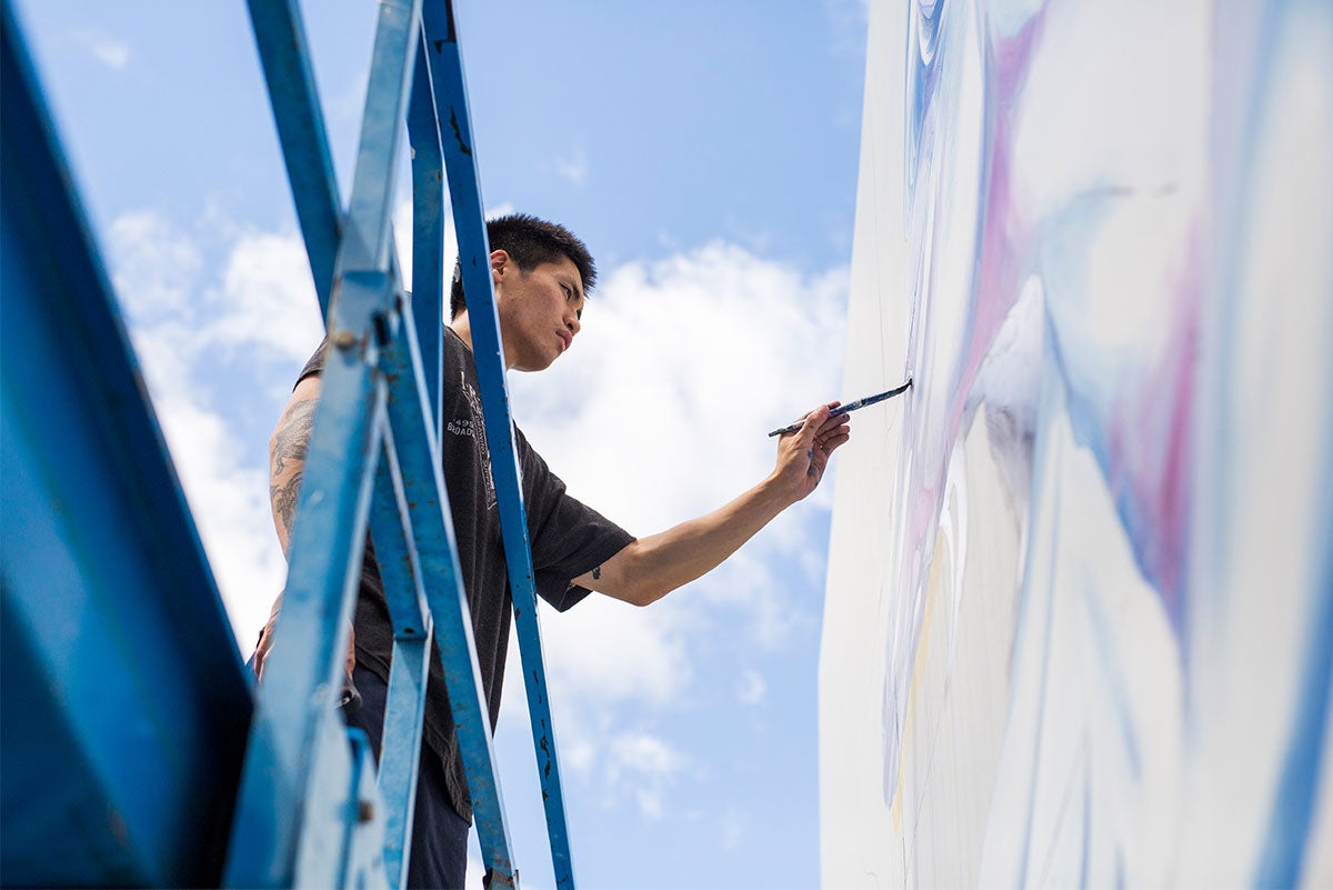 Shown at angle looking up from the ground, Boy Kong stands in a blue lift painting details on the mural.