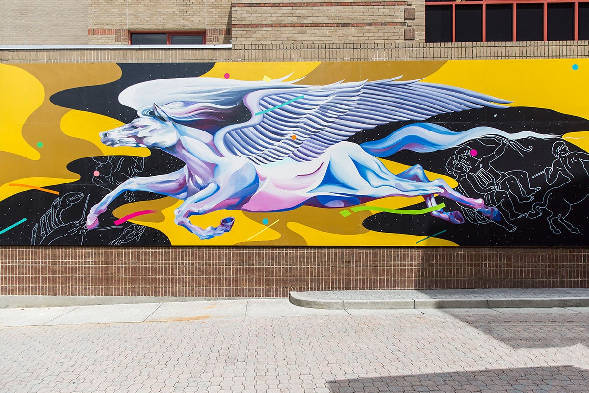 The completed Pegasus mural at UCF on the wall of the Student Union shows a white horse with giant wings, with shading done in pinks, purples, and blues, in flight on a yellow and black swirly background. In the black sections, you can see outlines of constellations. Neon colored dots and dashes in pinks, greens, and blues look like confetti.