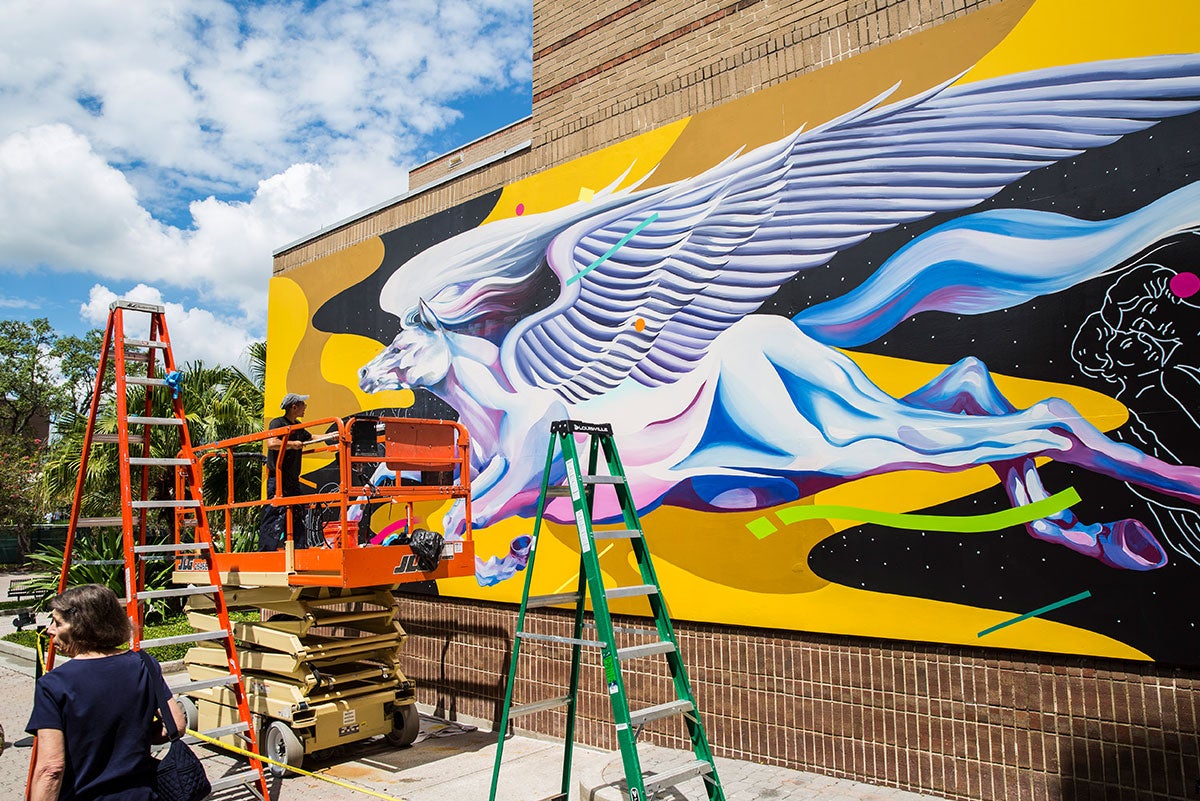 Viewed at an angle, the Pegasus mural at UCF is mostly complete. An orange lift and ladder and a green ladder are in front of it. In the lift are Boy Kong and Chris Troung. A woman is walking past.