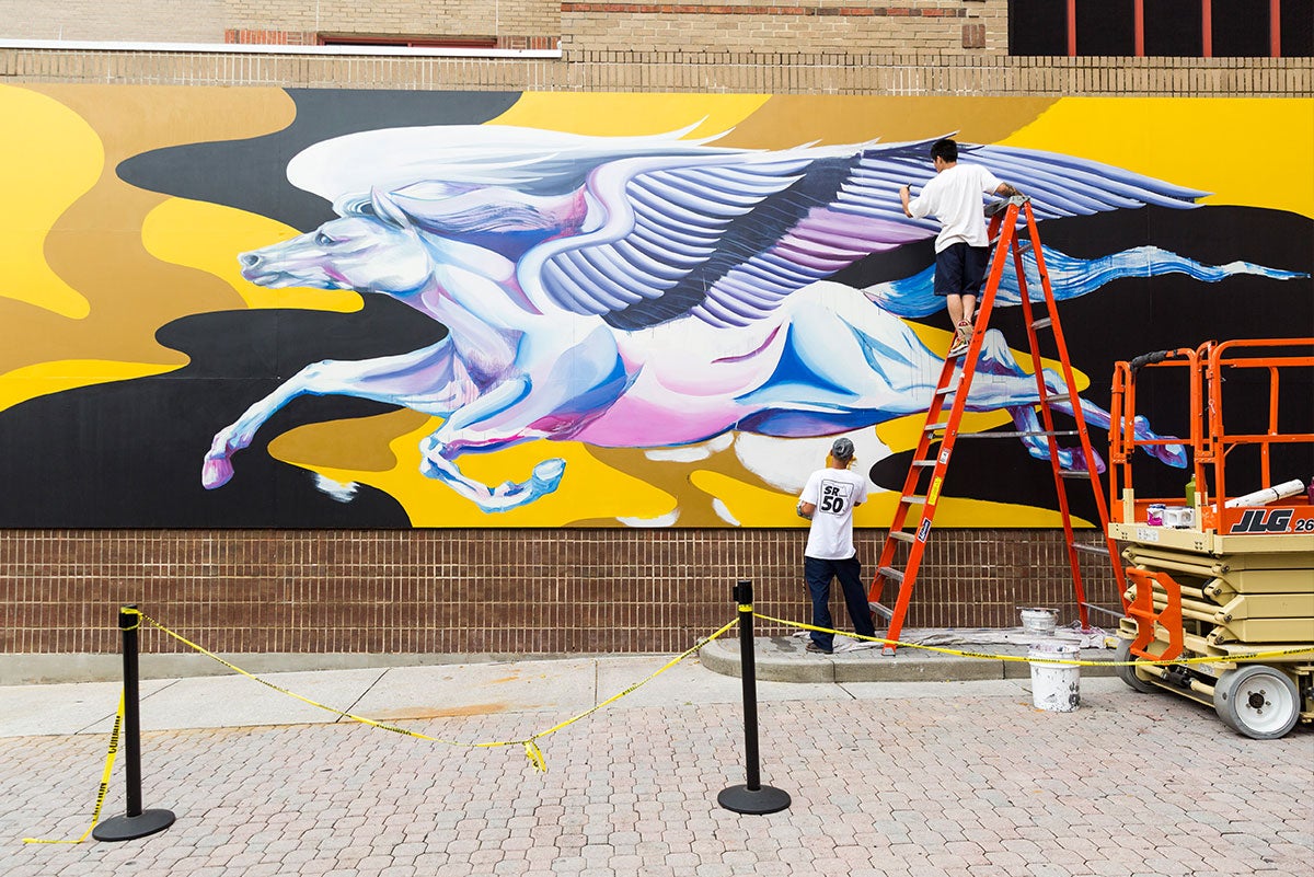 A wide shot of the mural at UCF shows a mostly completed horse with giant wings in flight on a black and yellow background. Boy Kong stands on top of an orange ladder in front of the mural, while Chris Troung holds it.