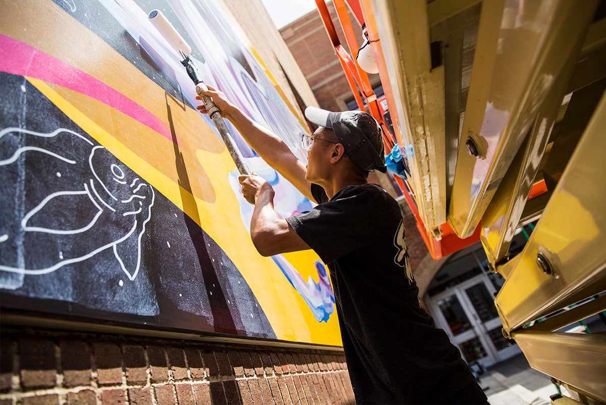 Standing between the mural and an orange lift, Chris Troung paints a clear top coat to the mural using a white roller.